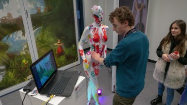 Photograph of someone playing the game The Body Keeps the Score, made for the jam. The game is about different kinds of touch and uses a mannequin with copper pads placed at various places on the body. In the photo, a player is touch the mannquin on its hip and looking at the attached laptop's screen.