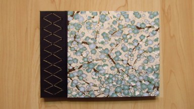 A view of the book's front cover. A vertical strip of black Japanese bookcloth, with decorative gold stitching forming a pattern of diamonds. The main front board is covered in Chiyogami paper, with cyan and white blossoms against dark branches.