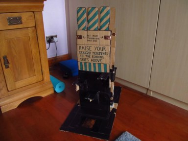 Photo of the cardboard tower the game revolves around, showing the blue player's side of the tower. There's a copper pad at the base of the tower, with 3 connection points staggered up the tower. Towards the top there are 2 copper pads, with the text 'PUT YOUR THUMBS HERE WHEN DONE'.