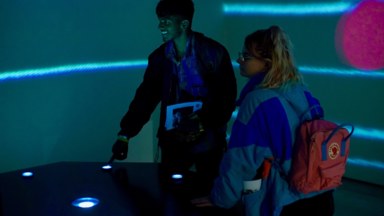 Two people stand behind the controller, interacting with it, lit by the buttons' white light and the projected visuals.