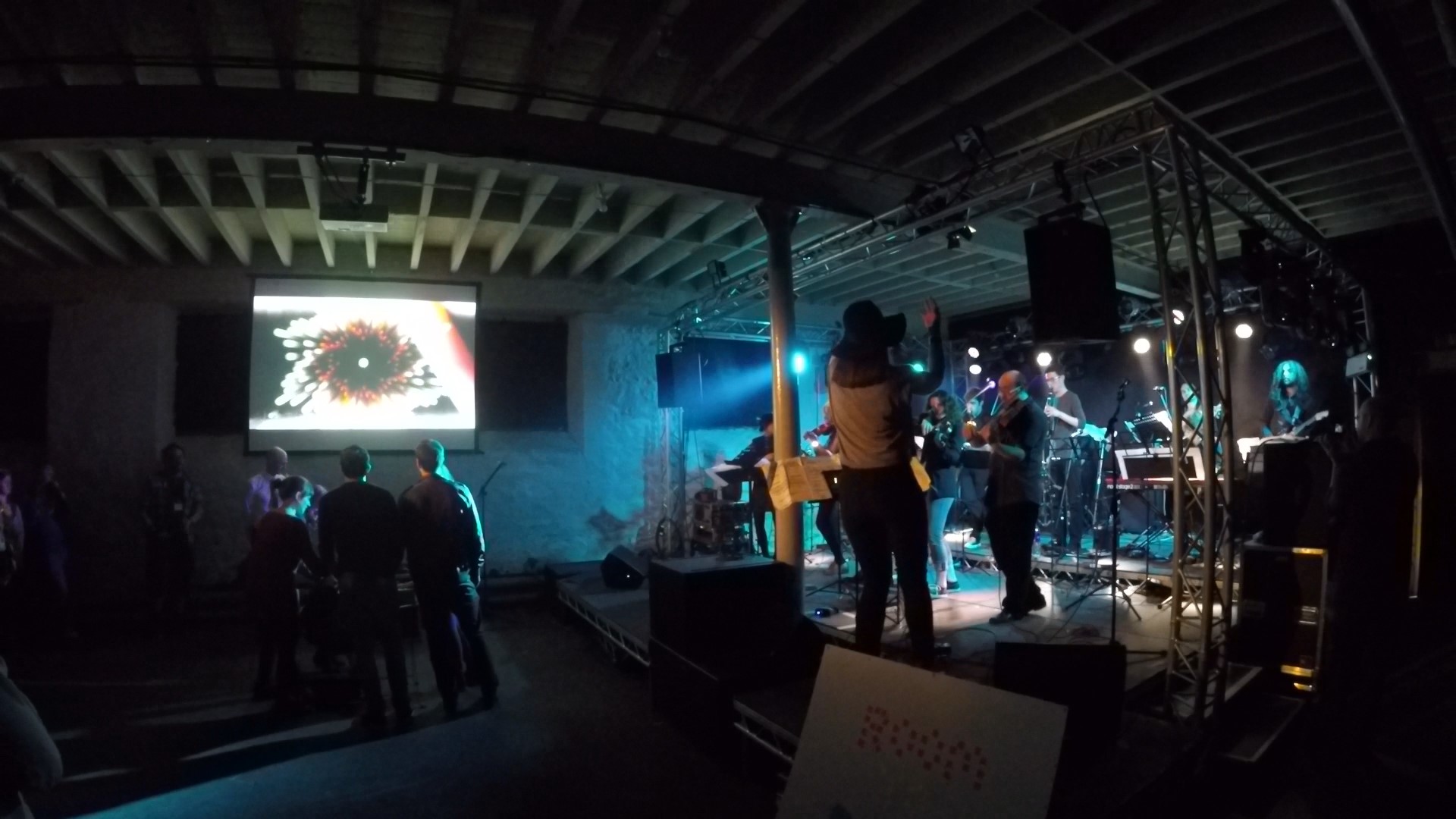 A shot of the event in action at Hidden Door 2016. The band are playing to the right, while Whispered Truths is visible on a projector to the left, with a handful of audience members clustered around the controller, manipulating the visuals and sound.