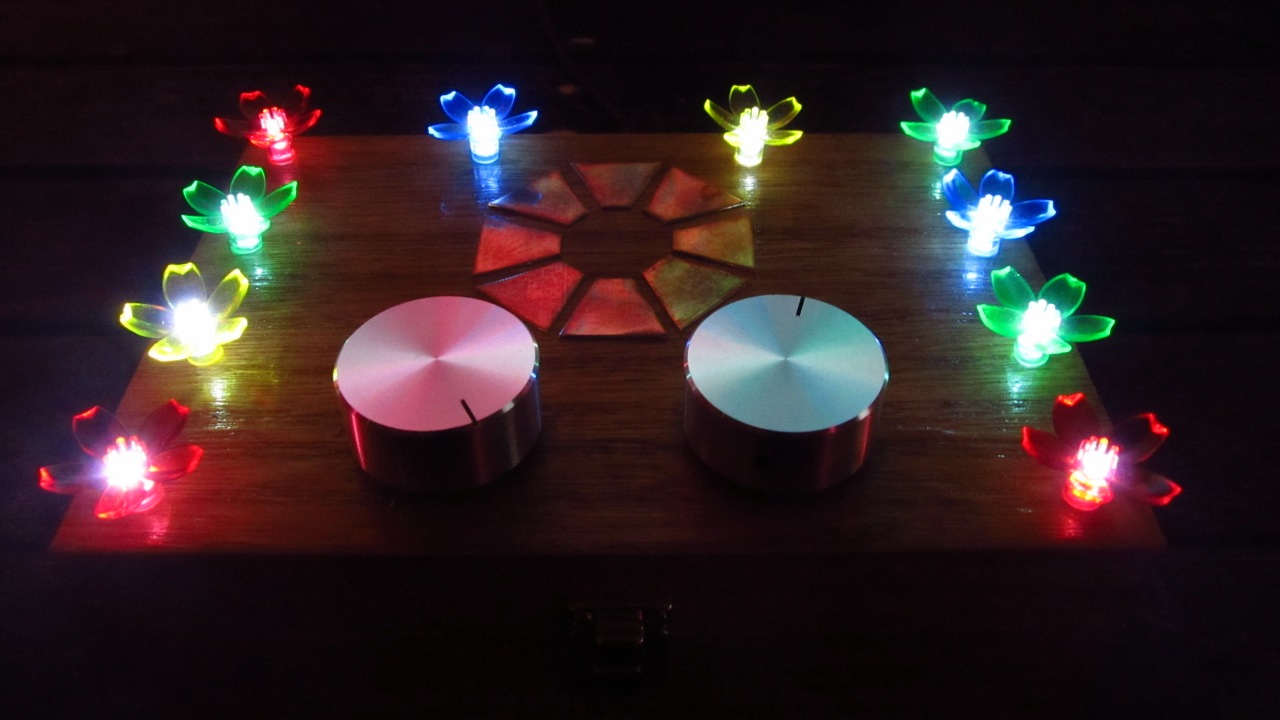Photo of the controller in the dark. There are 10 flower-shaped fairy lights arranged around the edges of a wooden tea box, with 2 large dials at the front, and an octagon of 8 brass plates arranged in the centre of the box.