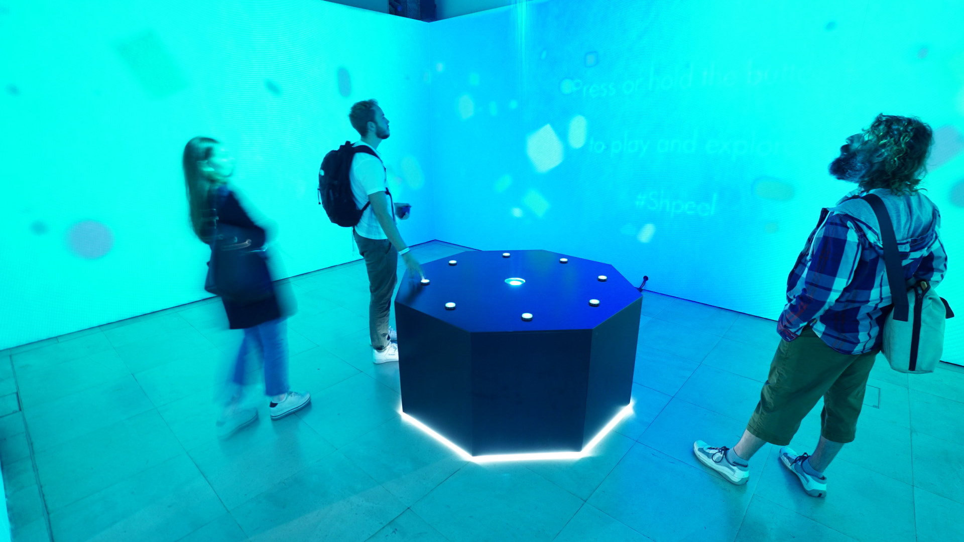 Photo of the installation in action. A large black octagon with 8 white illuminated buttons, and visuals projected onto all 4 surrounding walls. Three people stand around the controller, interacting with it.